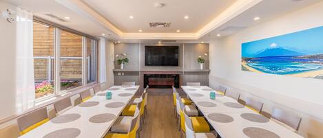 A spacious dining area with long tables and yellow chairs, a large TV screen, and a painting of a beach scene on the wall.