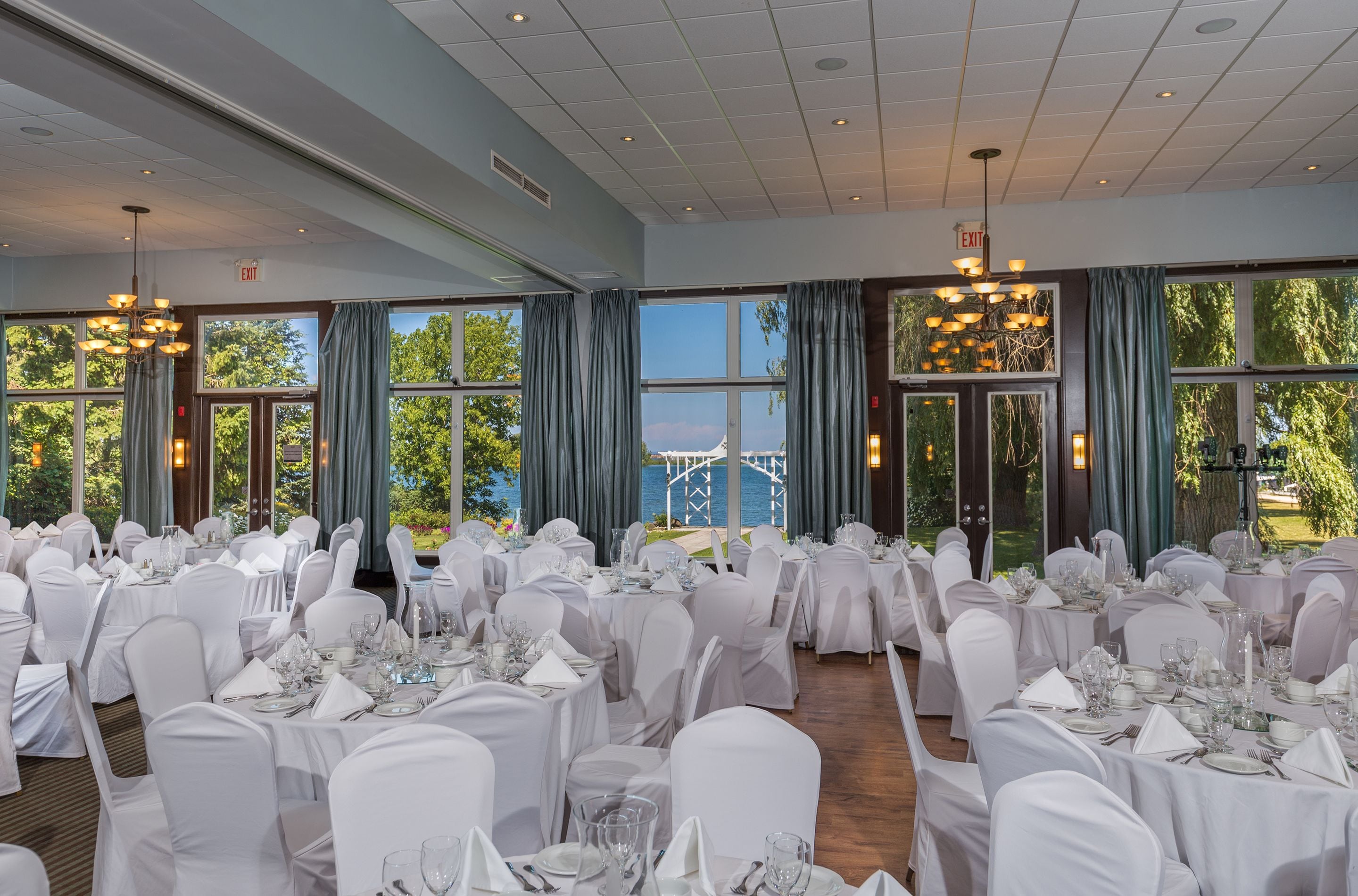A banquet hall with tables set for a formal event, large windows with curtains, and an exit sign.