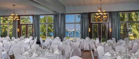 Un salón de banquetes con mesas dispuestas para un evento formal, ventanas amplias con cortinas y una señal de salida.