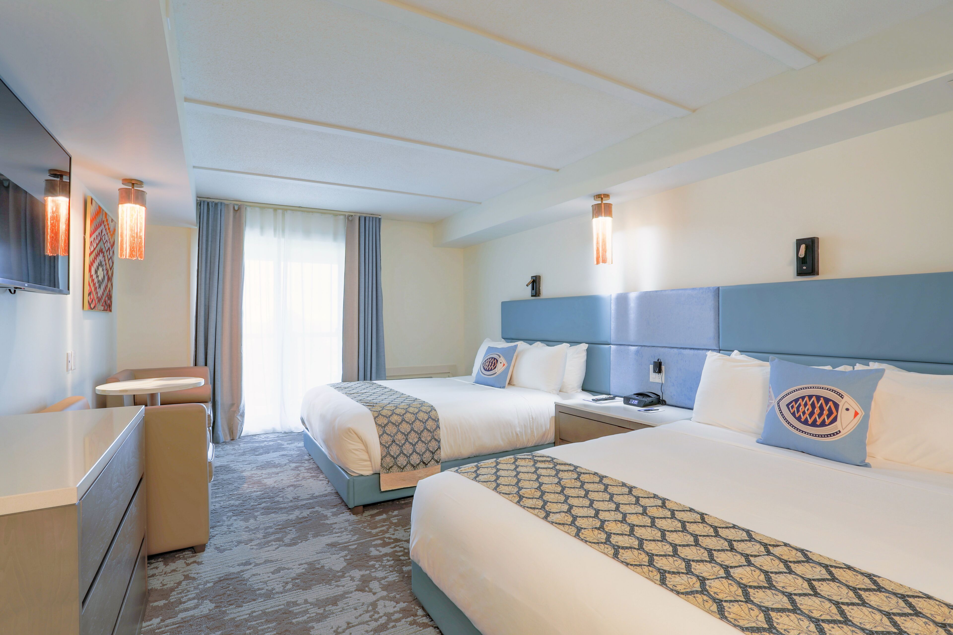 A hotel room with two beds, a desk, and a chair. There are two bedside tables with lamps, a window with curtains, and a wall-mounted phone.