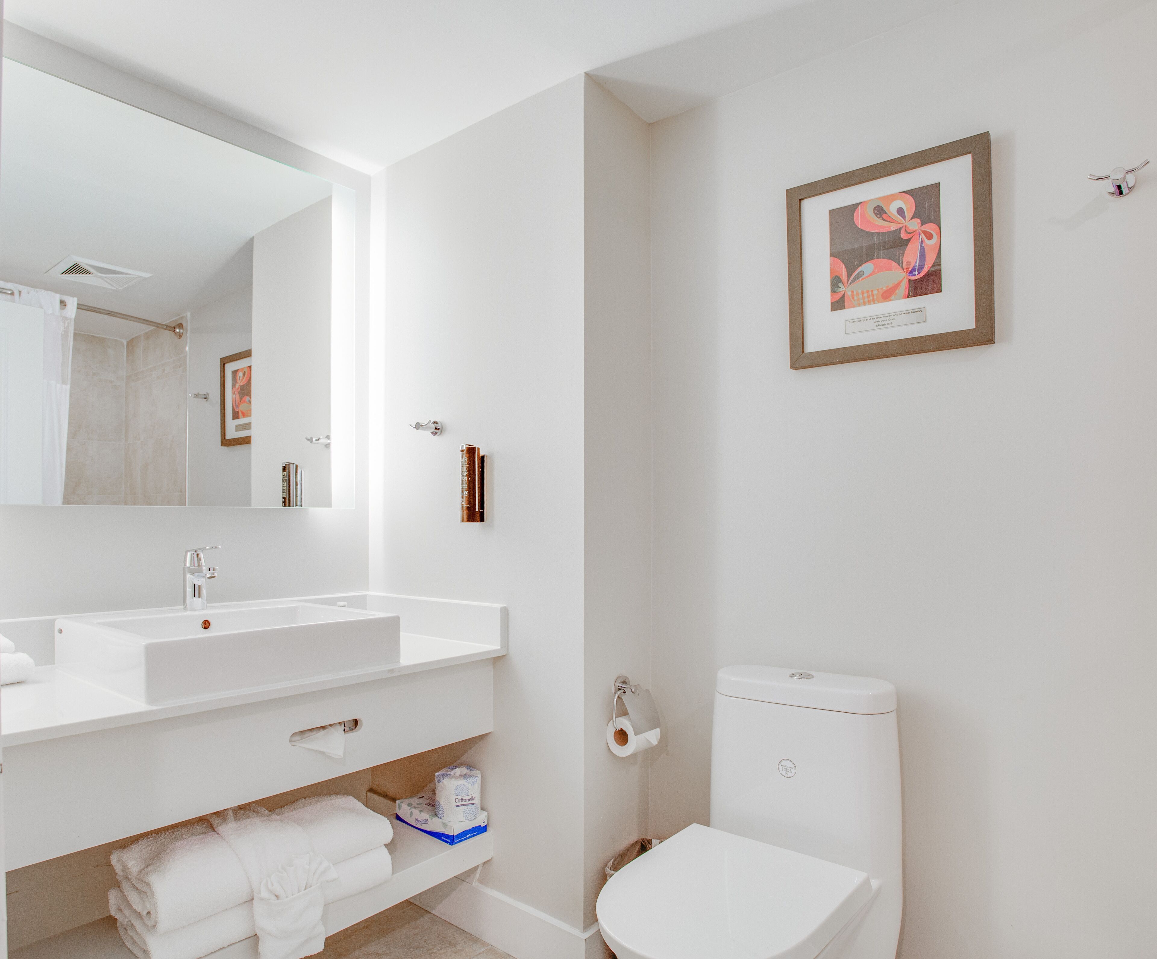 A modern bathroom with a white sink, mirror, and toilet. There is a framed artwork on the wall and a towel rack.