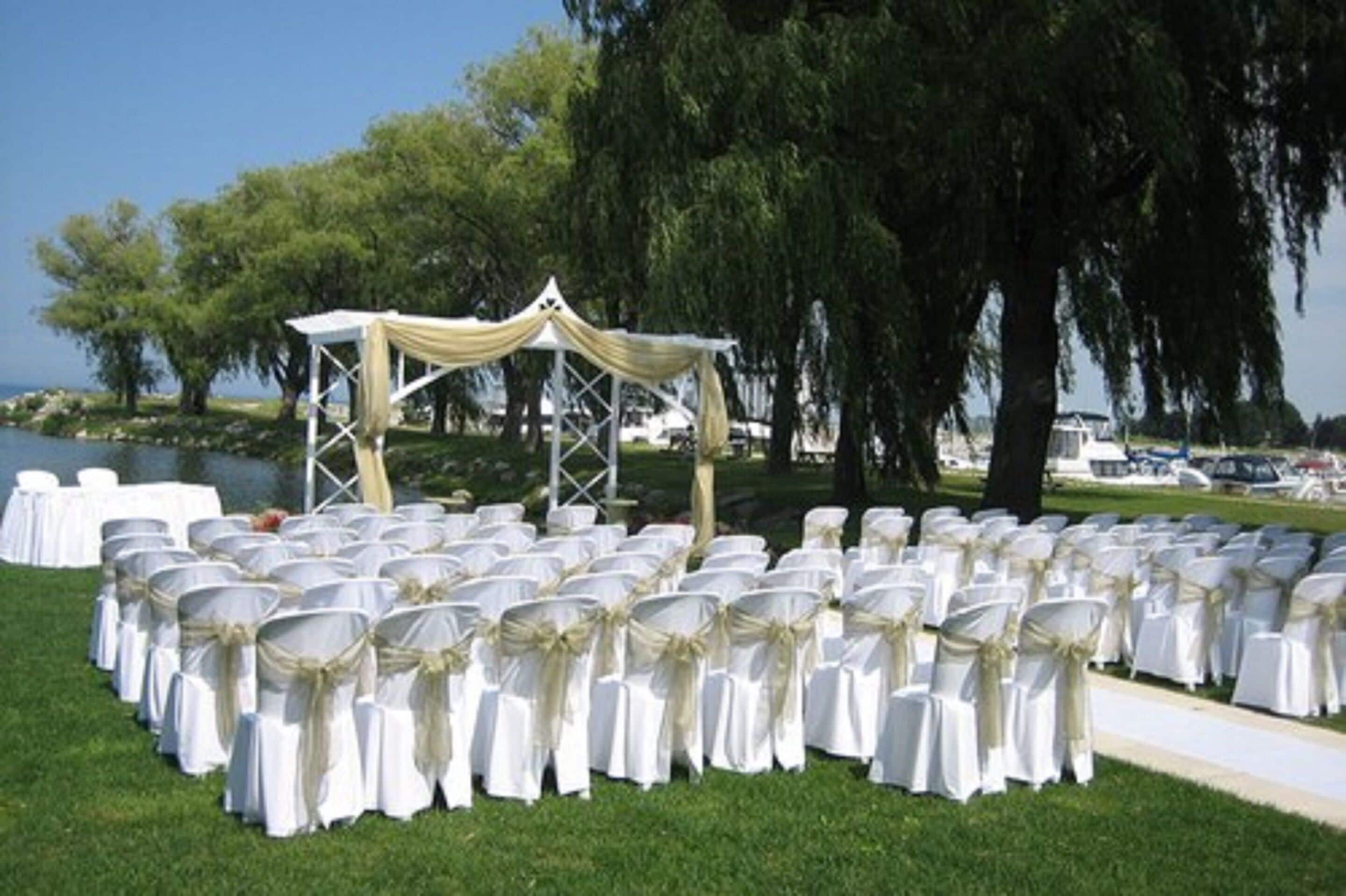 A wedding setup with rows of white chairs with gold sashes, a white canopy, and a body of water in the background.