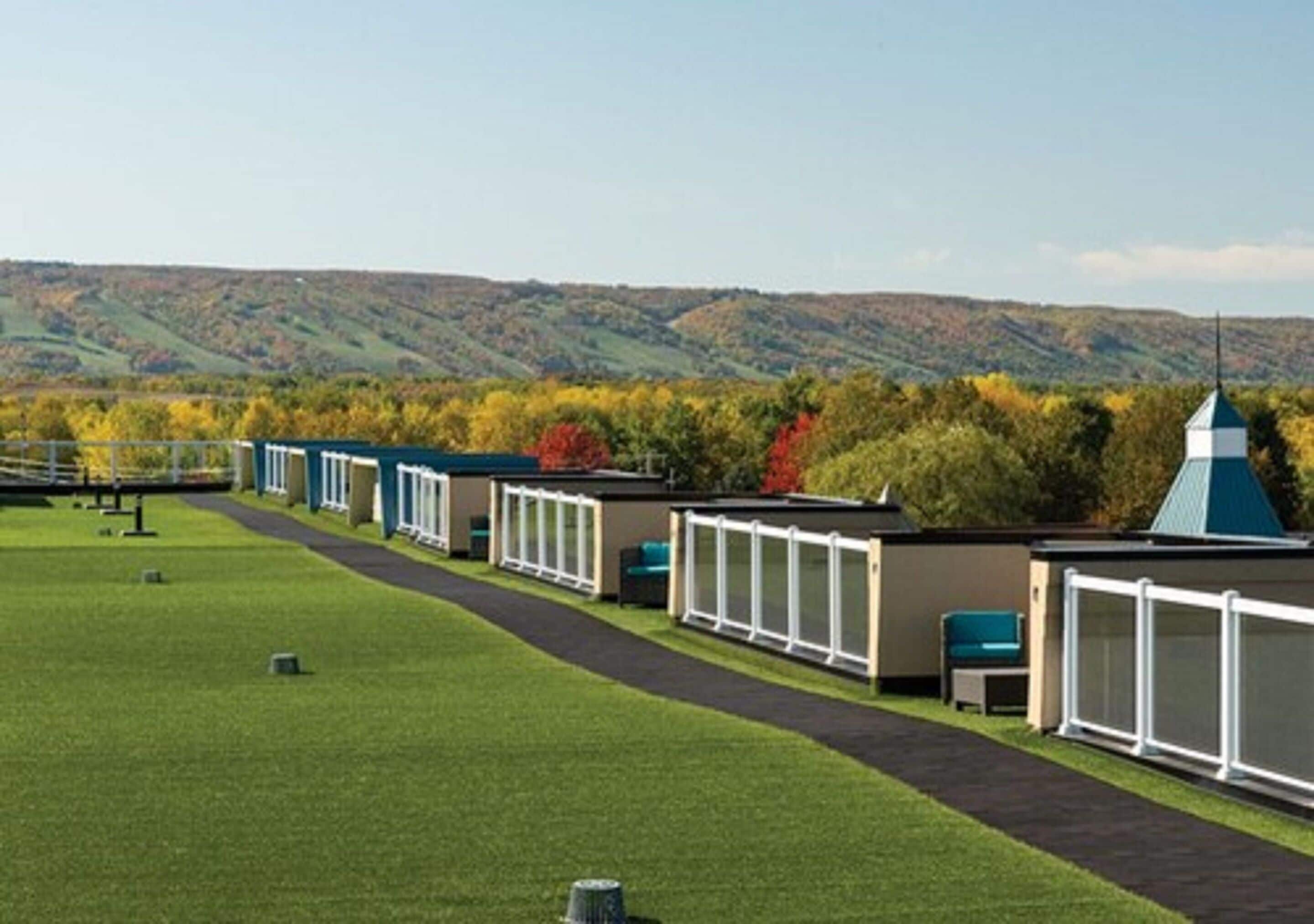 A row of cabins with a grassy path between them, set against a backdrop of autumn-colored trees and hills.