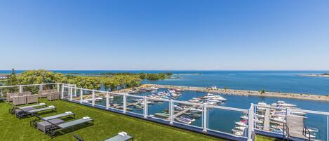 A rooftop terrace with lounge chairs and tables overlooking a marina with boats.