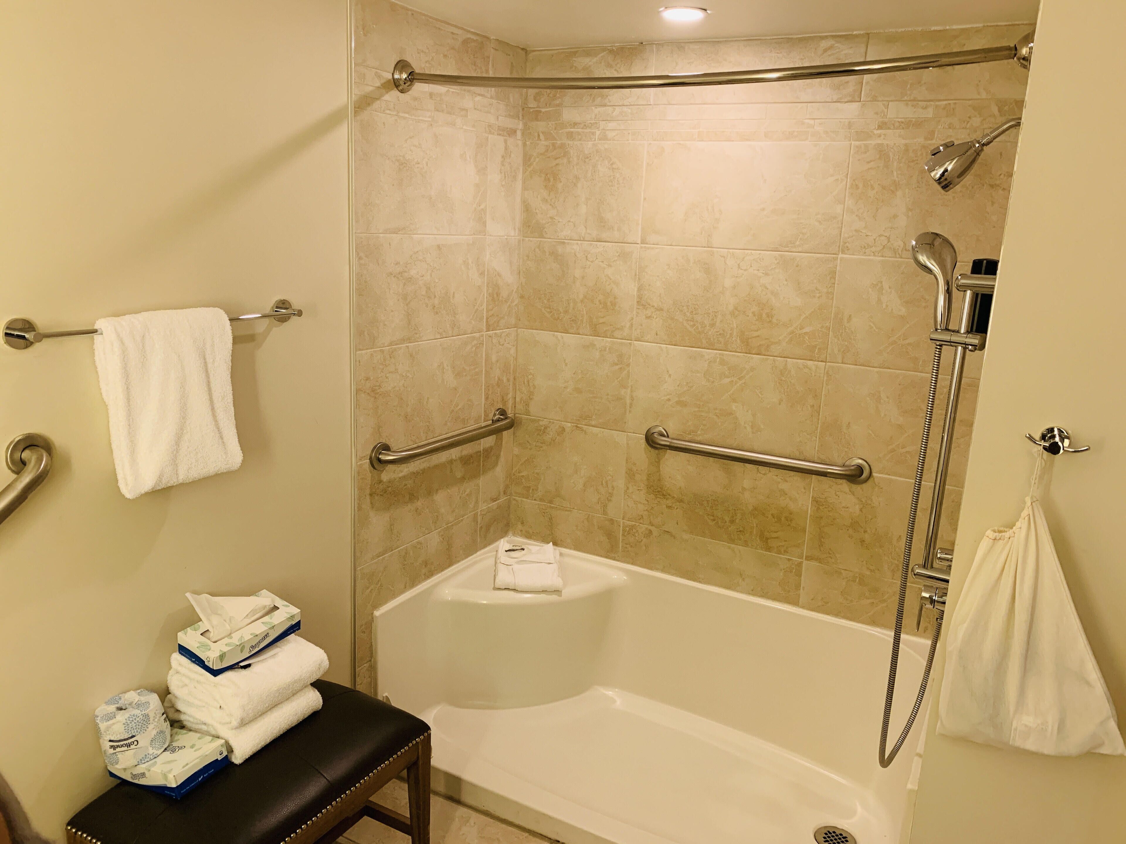 A bathroom with a bathtub, a shower, and a handrail. There are towels and toiletries on a bench.