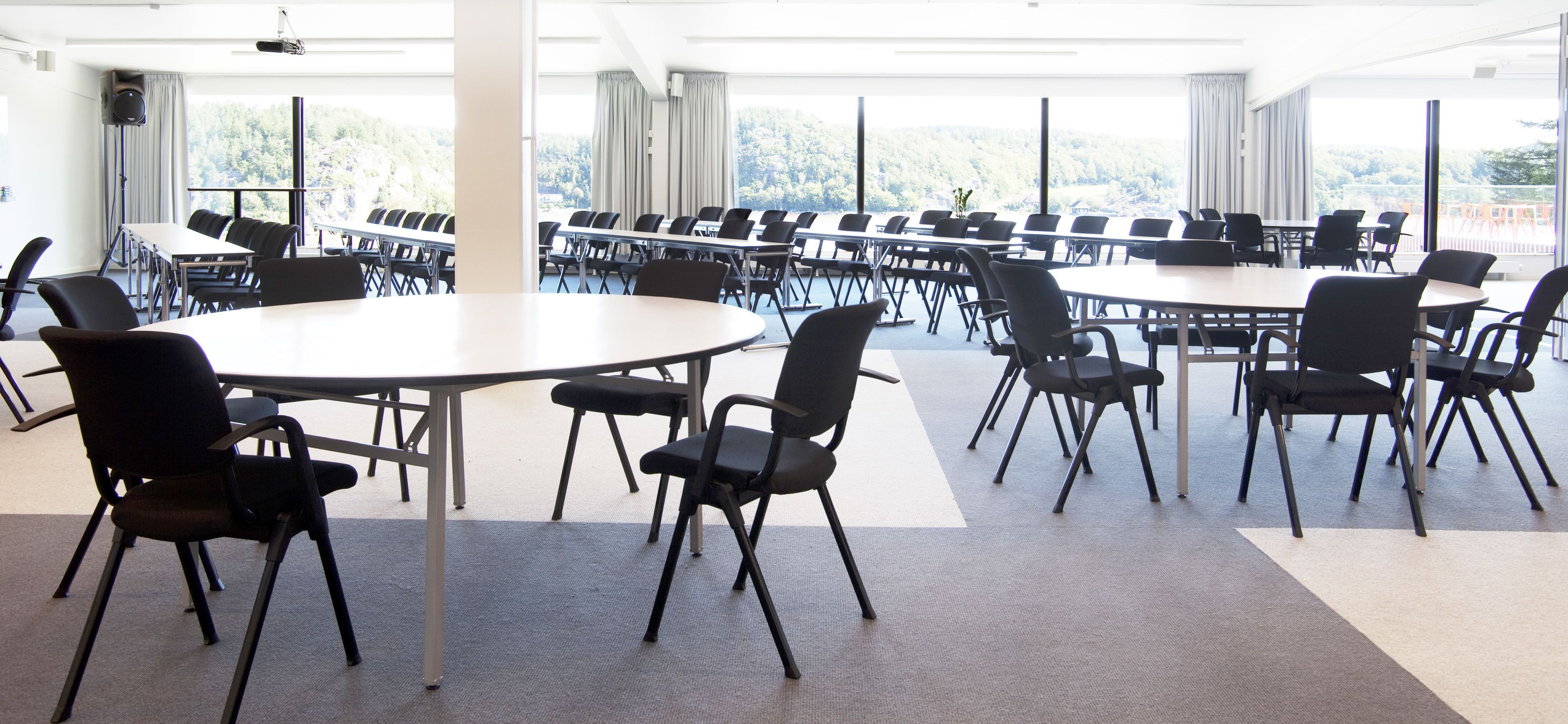Ein Konferenzraum mit mehreren Tischen und Stühlen, großen Fenstern mit Vorhängen sowie einer Sprechanlage.