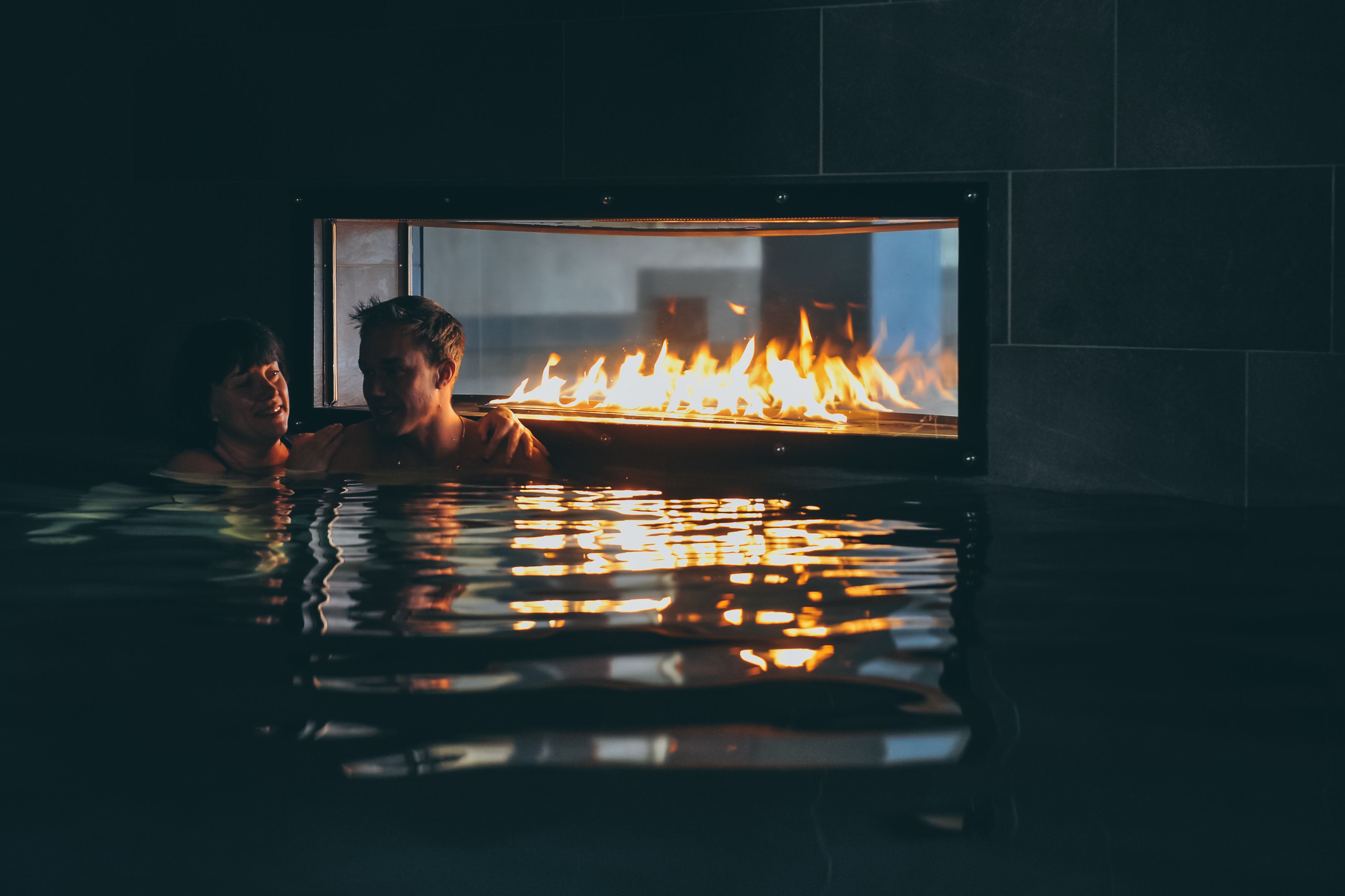 A couple in a heated indoor pool with a fire feature.