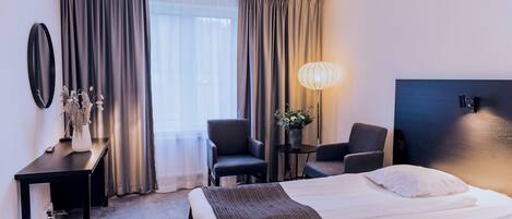 Habitación de hotel con cama, escritorio, dos sillas, lámpara y una ventana grande con cortinas.