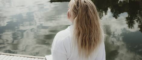 Una persona sentada en un muelle de madera con vista a un lago tranquilo, árboles y una estructura pequeña a lo lejos.