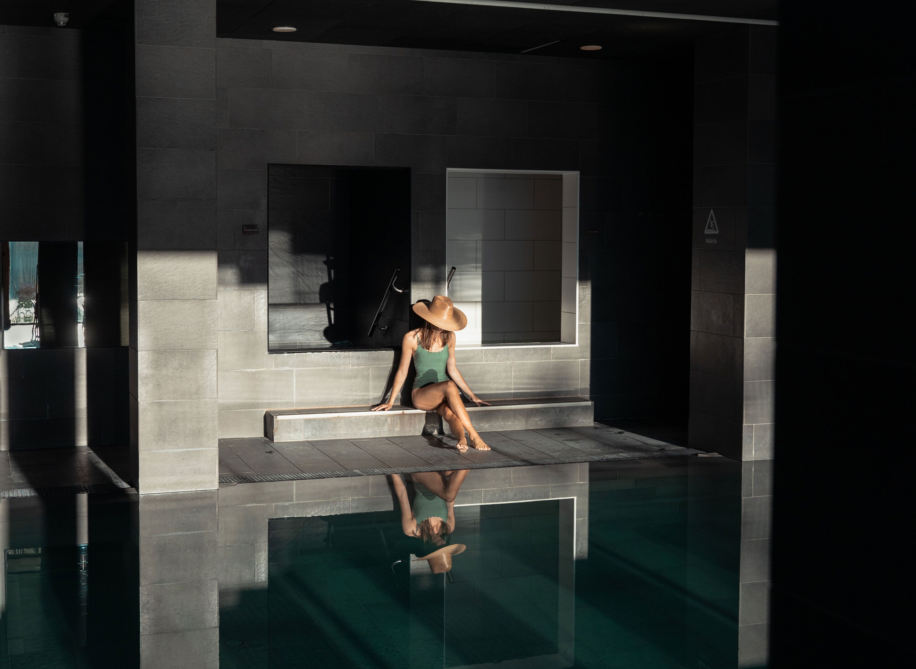 A woman sitting by a reflective pool in a modern building with large windows and a minimalist design.