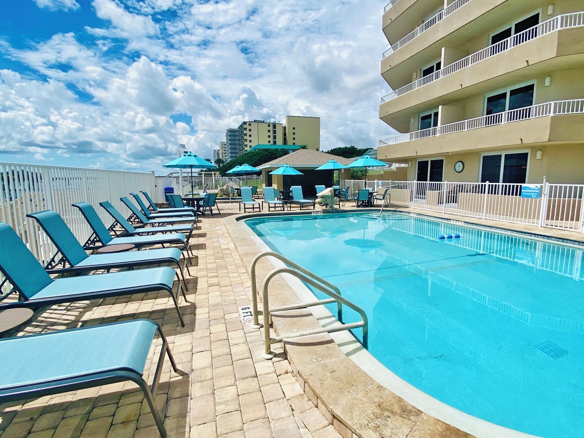 A poolside area with lounge chairs and umbrellas, a clear blue pool, and a white fence.