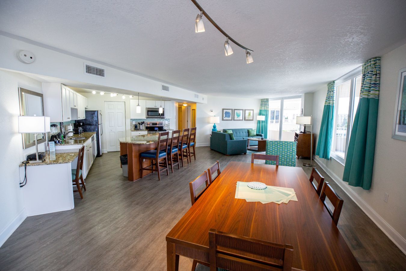 Een moderne keuken met witte kasten, een centraal eiland met barkrukken, een eethoek met een houten tafel en stoelen, en een zithoek met een bank en een televisie.