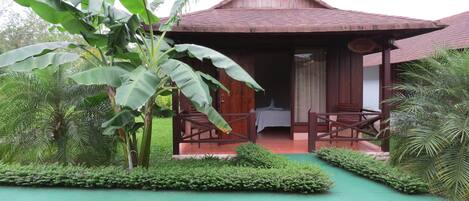 A wooden bungalow with a thatched roof, surrounded by lush greenery and a small pool.