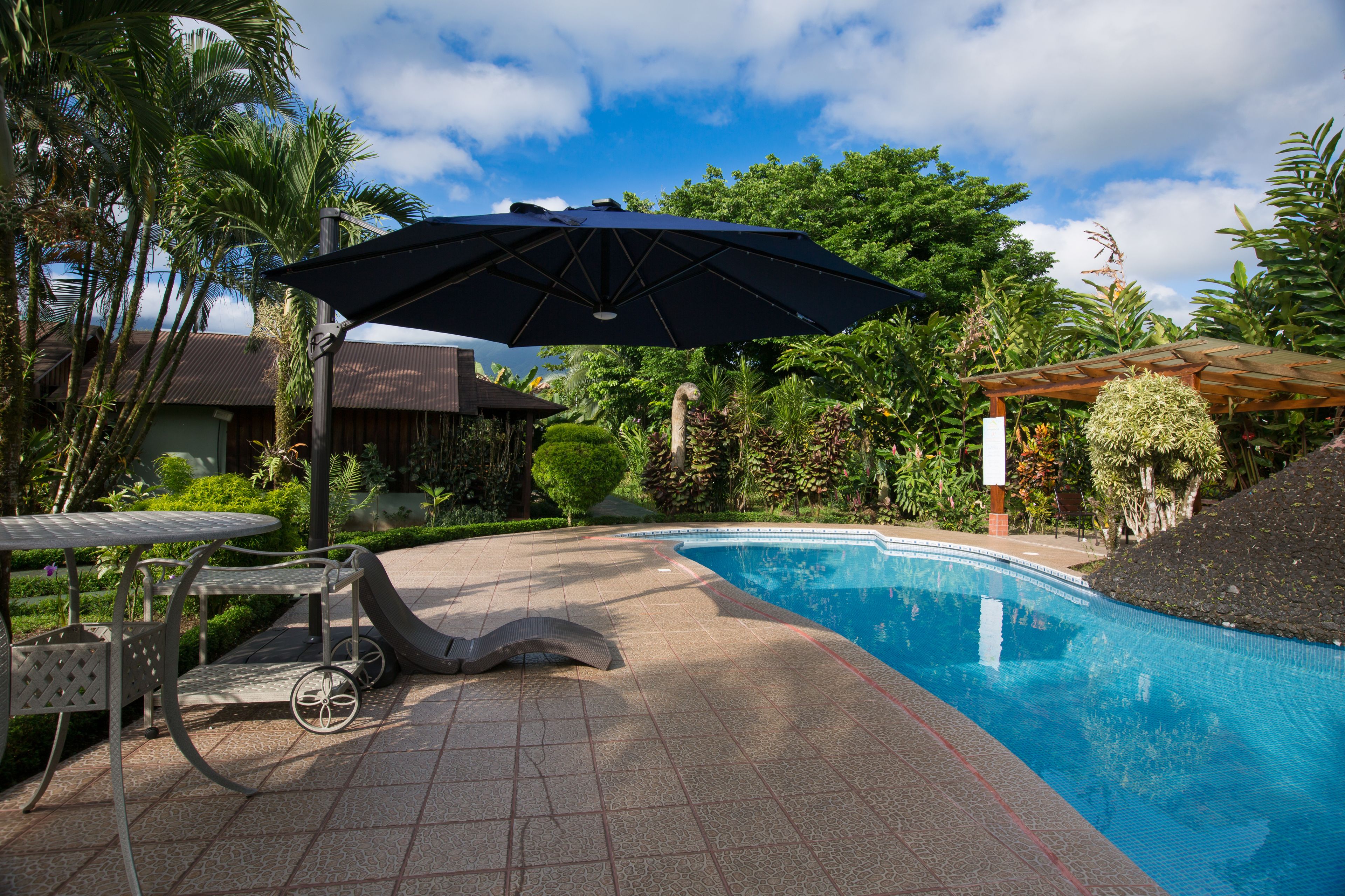 A swimming pool with a pergola and umbrella, surrounded by lush greenery and a tiled patio area.