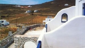 A white building with a stone wall and a blue door, overlooking a hilly landscape with other white buildings. - Piliouris Studios (Paros)