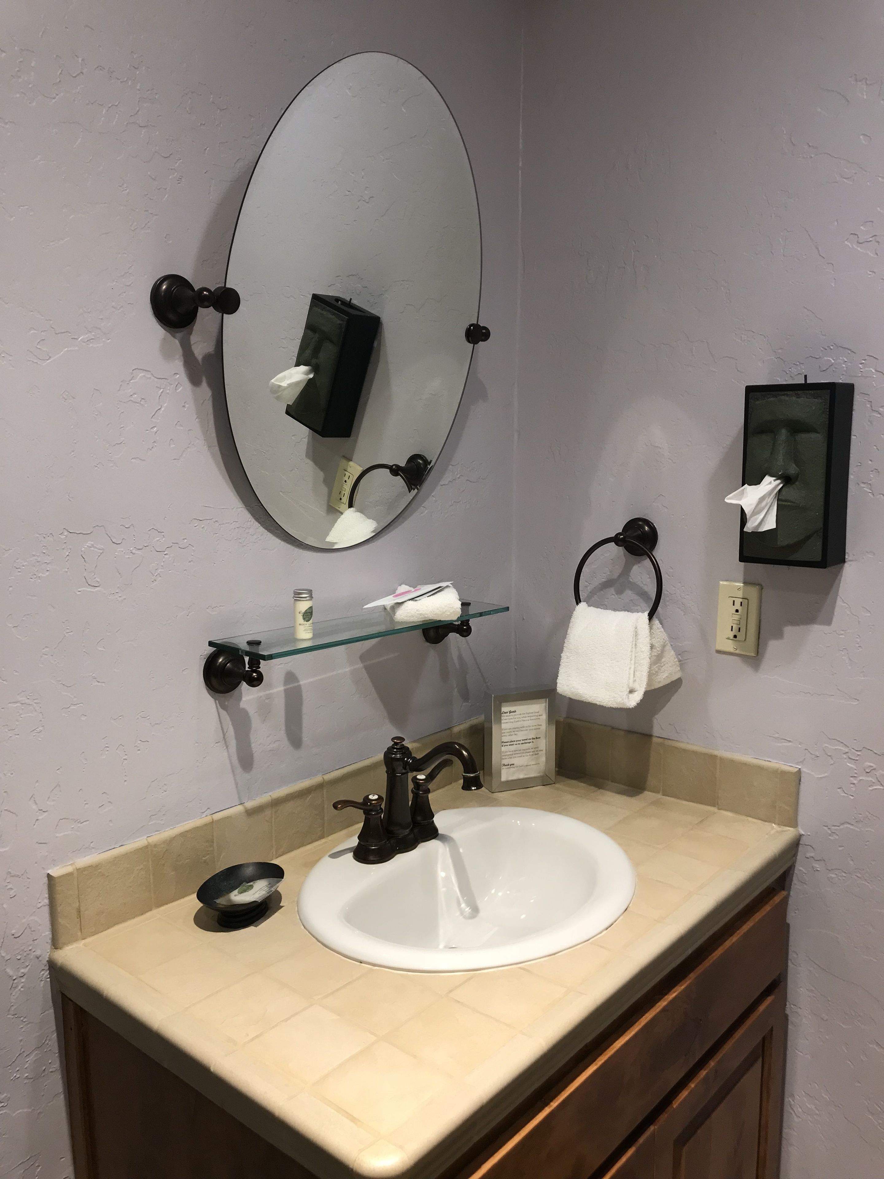 Une salle de bain avec un lavabo blanc, un miroir rond, un porte-serviettes, une étagère en verre et un panneau encadré.