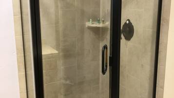 A glass-enclosed shower with a black frame, beige tiles, and a small shelf with toiletries.