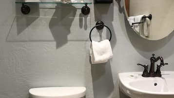 A bathroom with a toilet, sink, and mirror. There is a glass shelf above the toilet holding a framed picture and a tissue box.