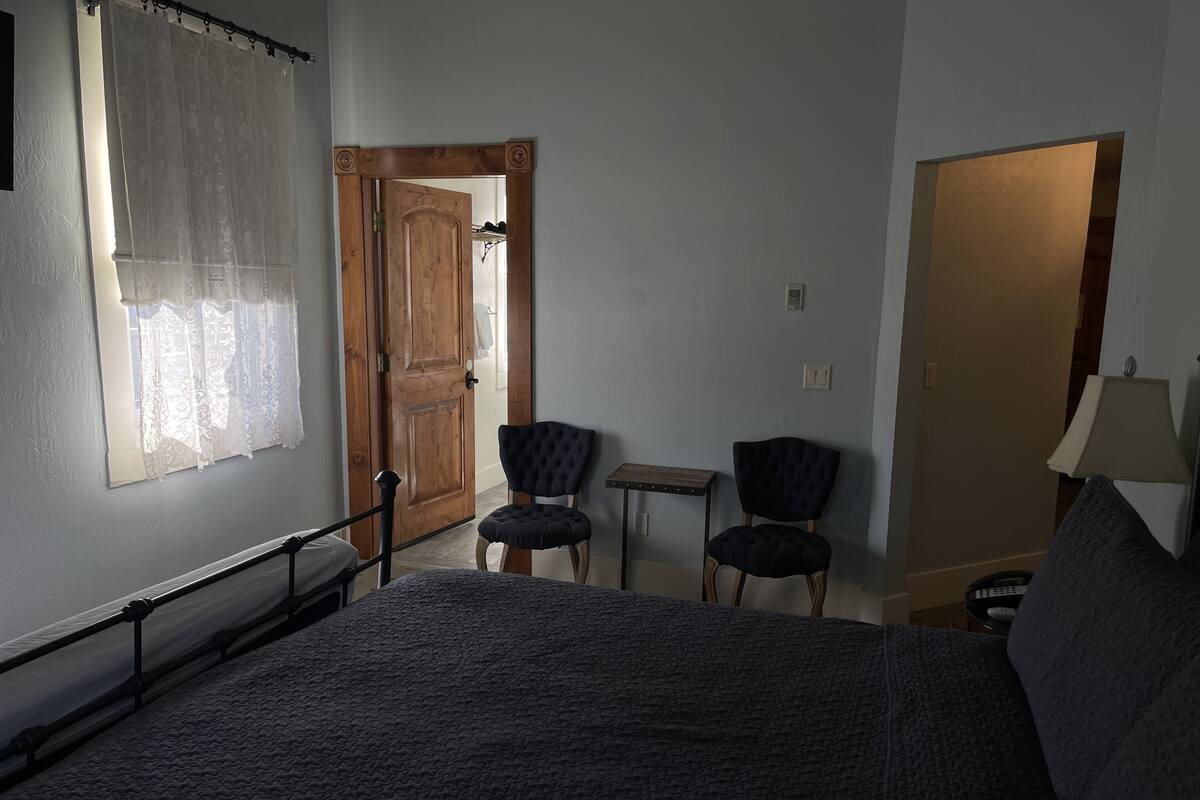 A bedroom with a bed, two chairs, a small table, a lamp, and a door leading to another room.