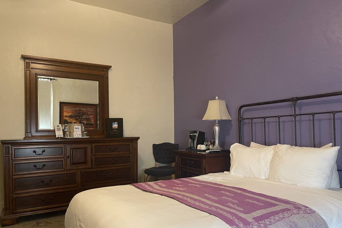 A hotel room with a large bed, a dresser with drawers, a nightstand with a lamp, and a chair.