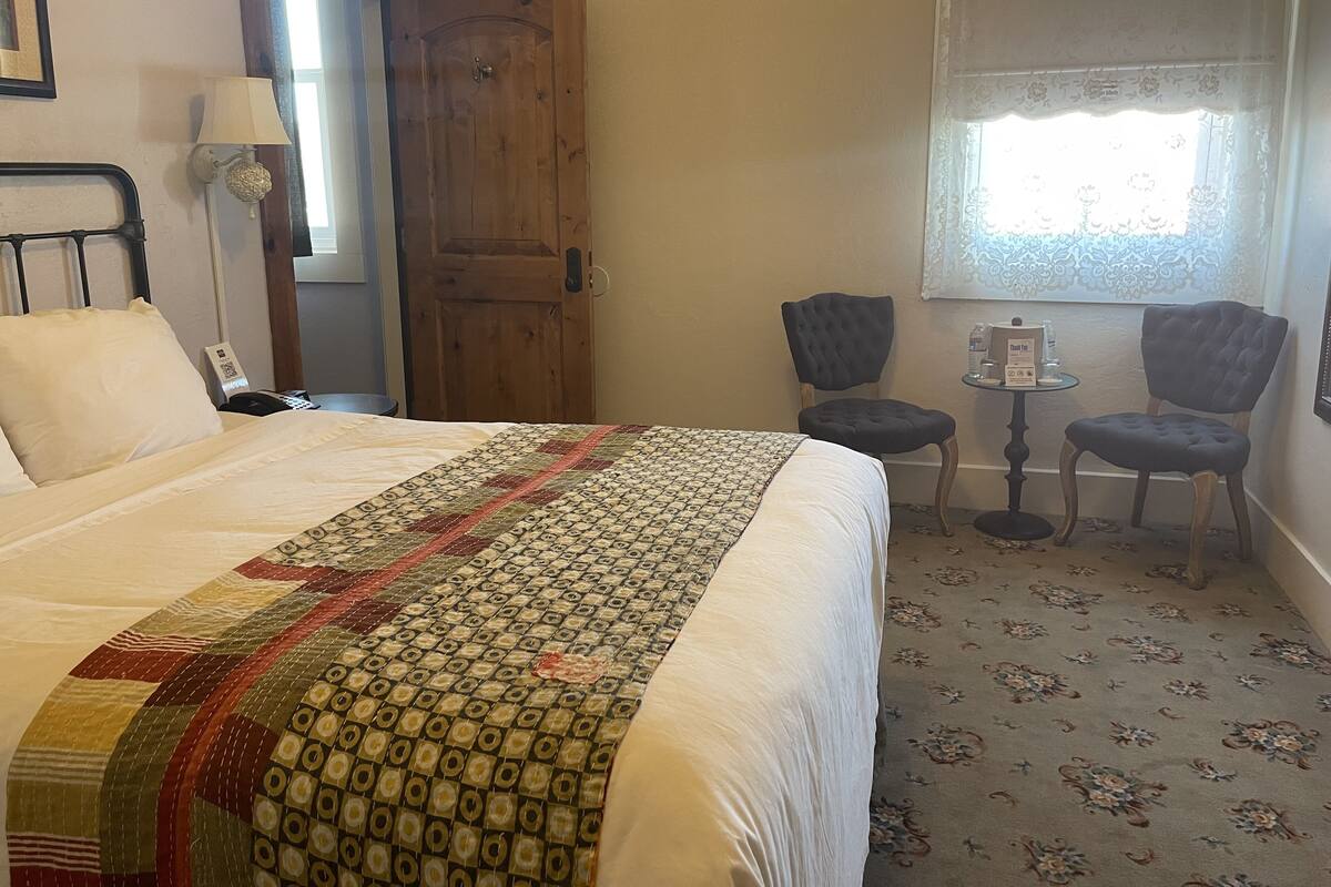A hotel room with a large bed, a wooden door, two upholstered chairs, a small table, and a window with a patterned curtain.