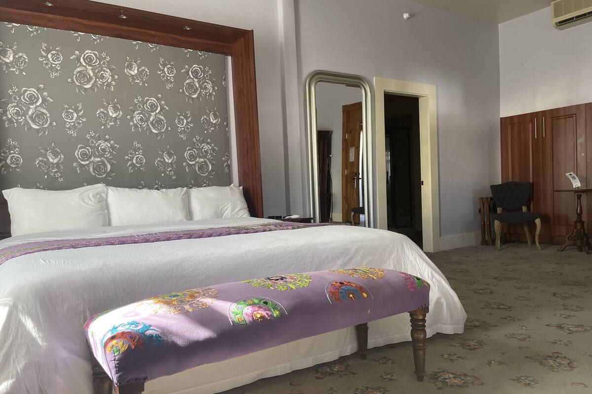 A bedroom with a large bed, a bench with colorful embroidery, a mirror, and a wooden wardrobe.