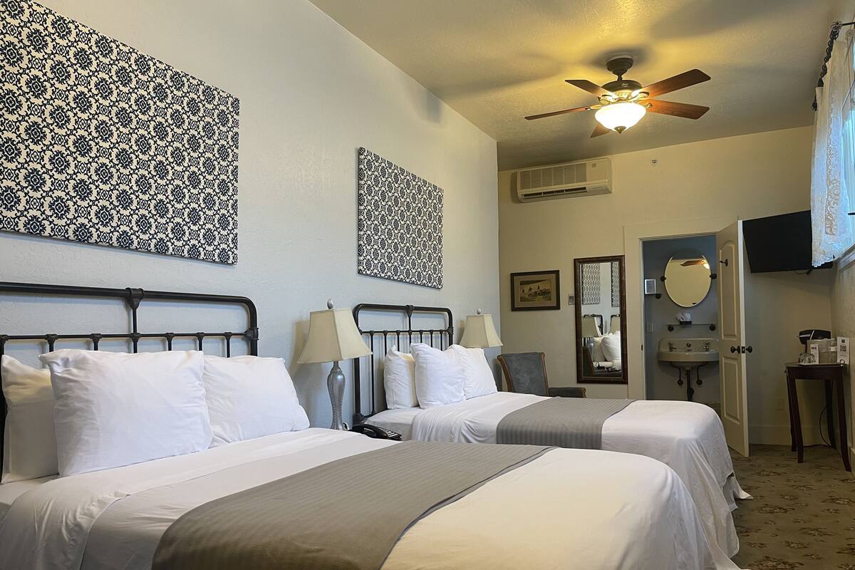 A hotel room with two beds, a ceiling fan, a mirror, a sink, and a television.