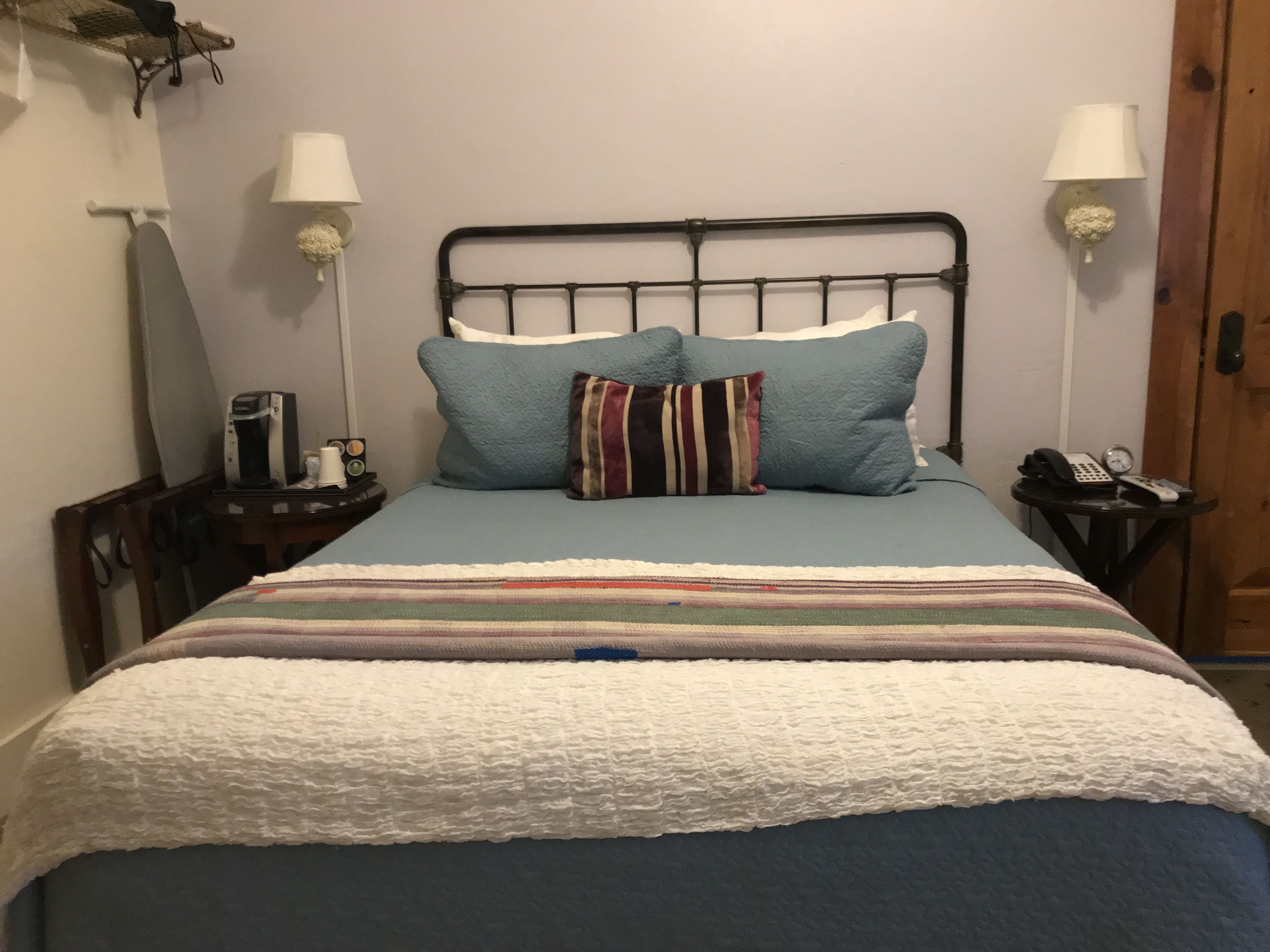 Een bed met een gestreepte deken, twee kussens, een metalen hoofdbord, nachtlampjes, een klok en een klein tafeltje met een telefoon en een afstandsbediening.