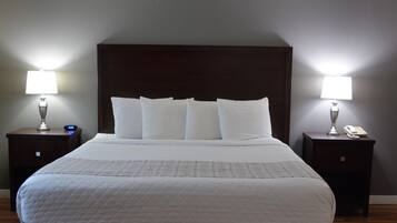 Una cama bien tendida con sábanas blancas y un cabecero oscuro. Hay dos mesitas de noche, cada una con una lámpara y un teléfono. El cuarto tiene piso de madera y paredes gris claro.