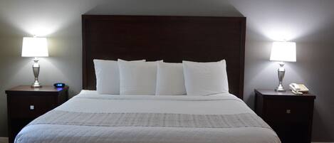 Een net opgemaakt bed met witte lakens en een donker hoofdbord. Twee nachtkastjes, elk met een lamp en een telefoon. De kamer heeft een houten vloer en lichtgrijze muren.