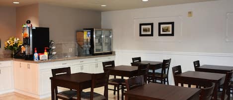 A dining area with wooden tables and chairs, a kitchen area with a refrigerator and microwave, and two framed pictures on the wall.