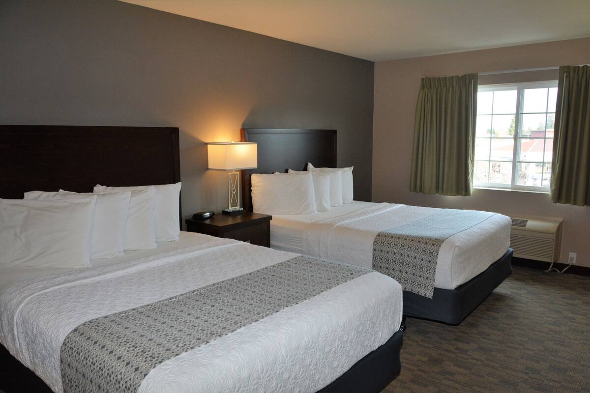 A hotel room with two beds, a nightstand with a lamp, a window with curtains, and a wall-mounted air conditioner.