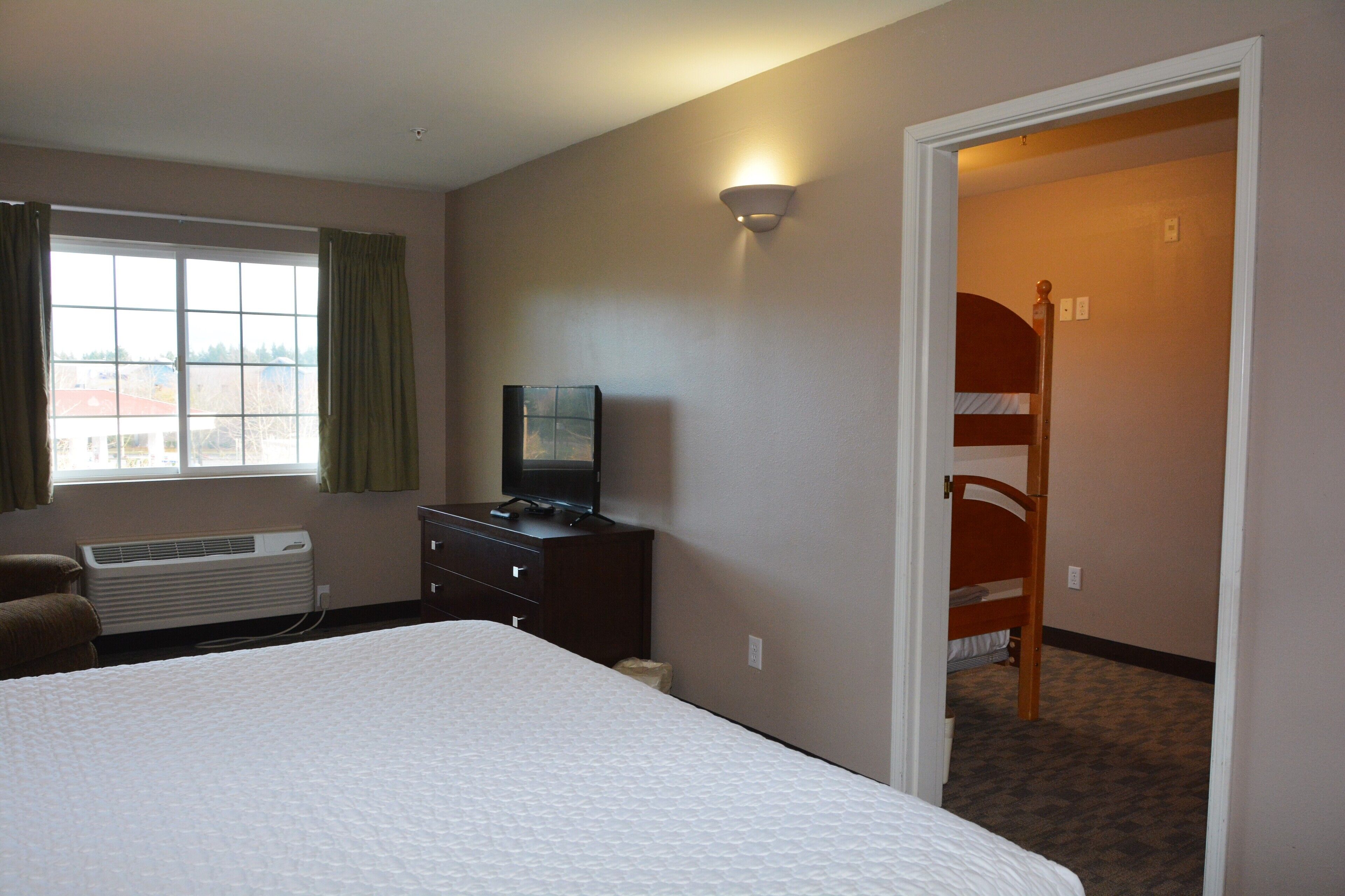 Une chambre d’hôtel avec un grand lit, un téléviseur posé sur une commode, une fenêtre avec des rideaux et un lit superposé dans un coin.