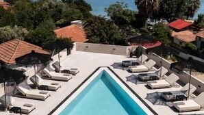 Una piscina rectangular con sillones de descanso y sombrillas, rodeada de vegetación y edificios con techos de tejas rojas, con vistas a una masa de agua y una isla distante.