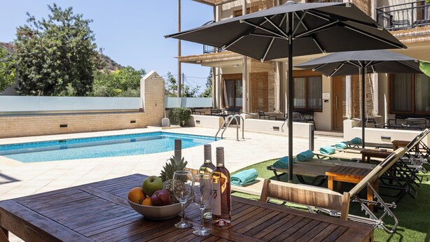 A poolside area with a wooden table, lounge chairs, and umbrellas. - Pantheon Villas & Suites  (Mylopotamos)