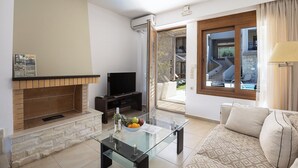 A modern living room with a glass coffee table, a flat-screen TV, a stone fireplace, and a view of the outdoor area. - Pantheon Villas & Suites  (Mylopotamos)