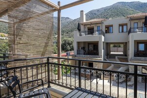 Un balcón con vistas a un edificio de dos pisos, mobiliario exterior y un paisaje montañoso.