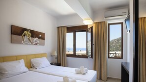A hotel room with two beds, a painting on the wall, a window with a view, and an air conditioning unit. - Pantheon Villas & Suites  (Mylopotamos)