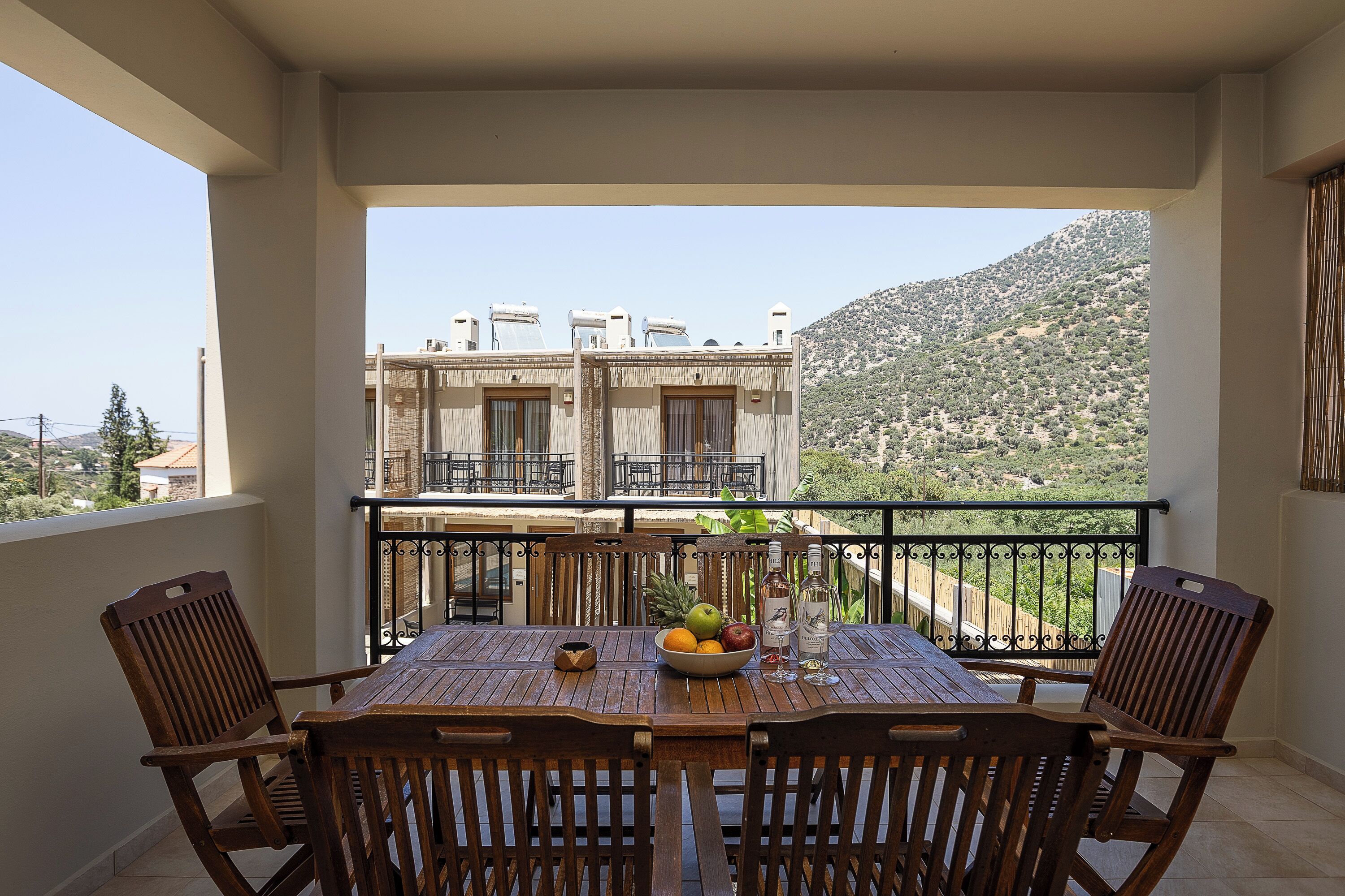 Un balcon avec une table et des chaises en bois, un saladier rempli de fruits et une bouteille d’eau.