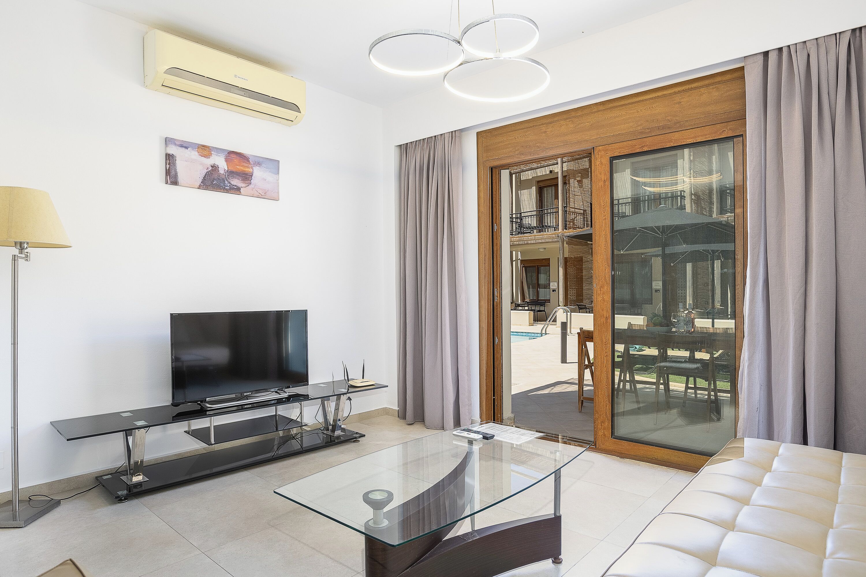 A modern living room with a flat-screen TV, glass coffee table, and a sofa. There is a sliding glass door leading to an outdoor area with a pool and furniture.
