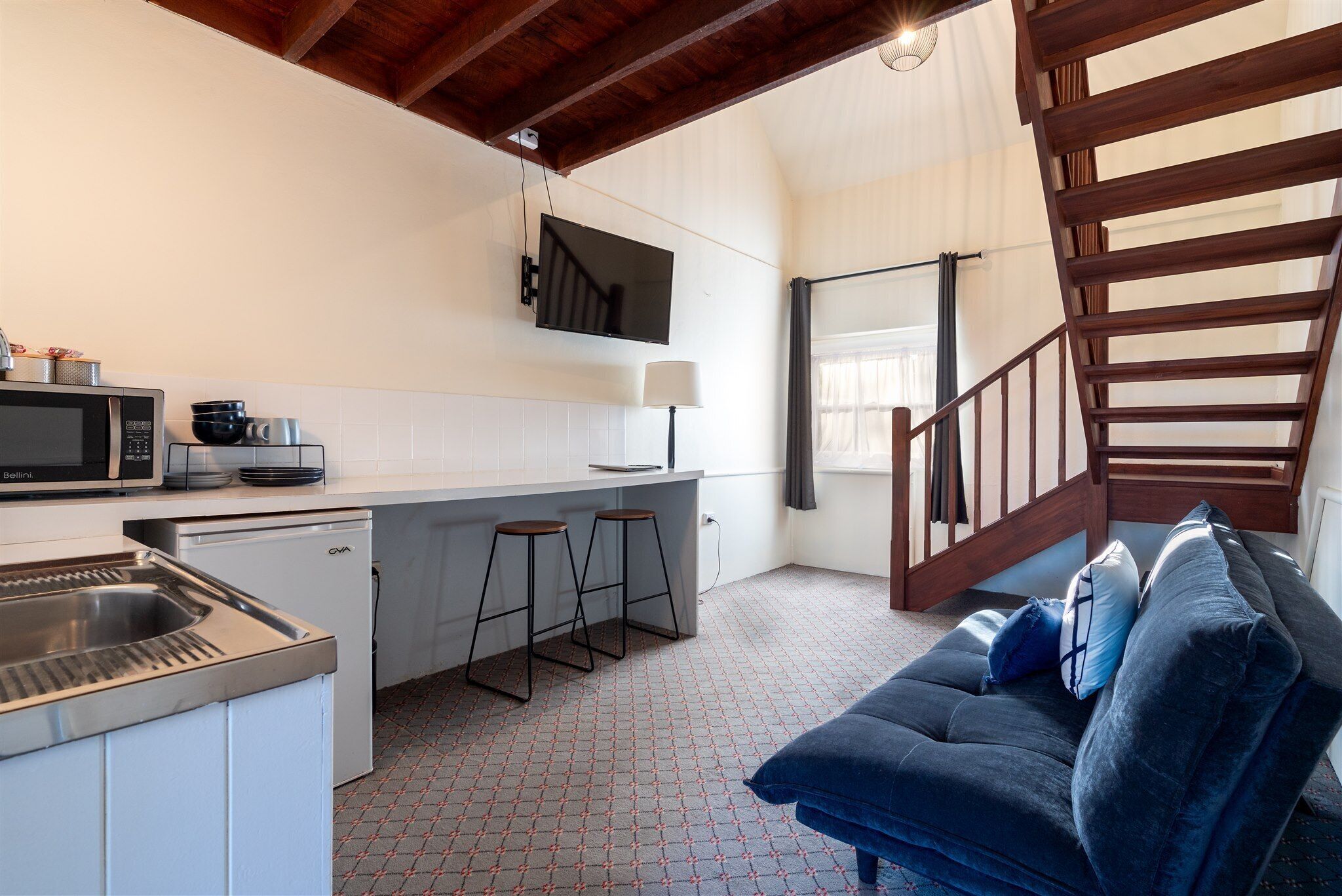 A compact kitchen with a microwave, sink, and stools, a sofa with cushions, and a wooden staircase.