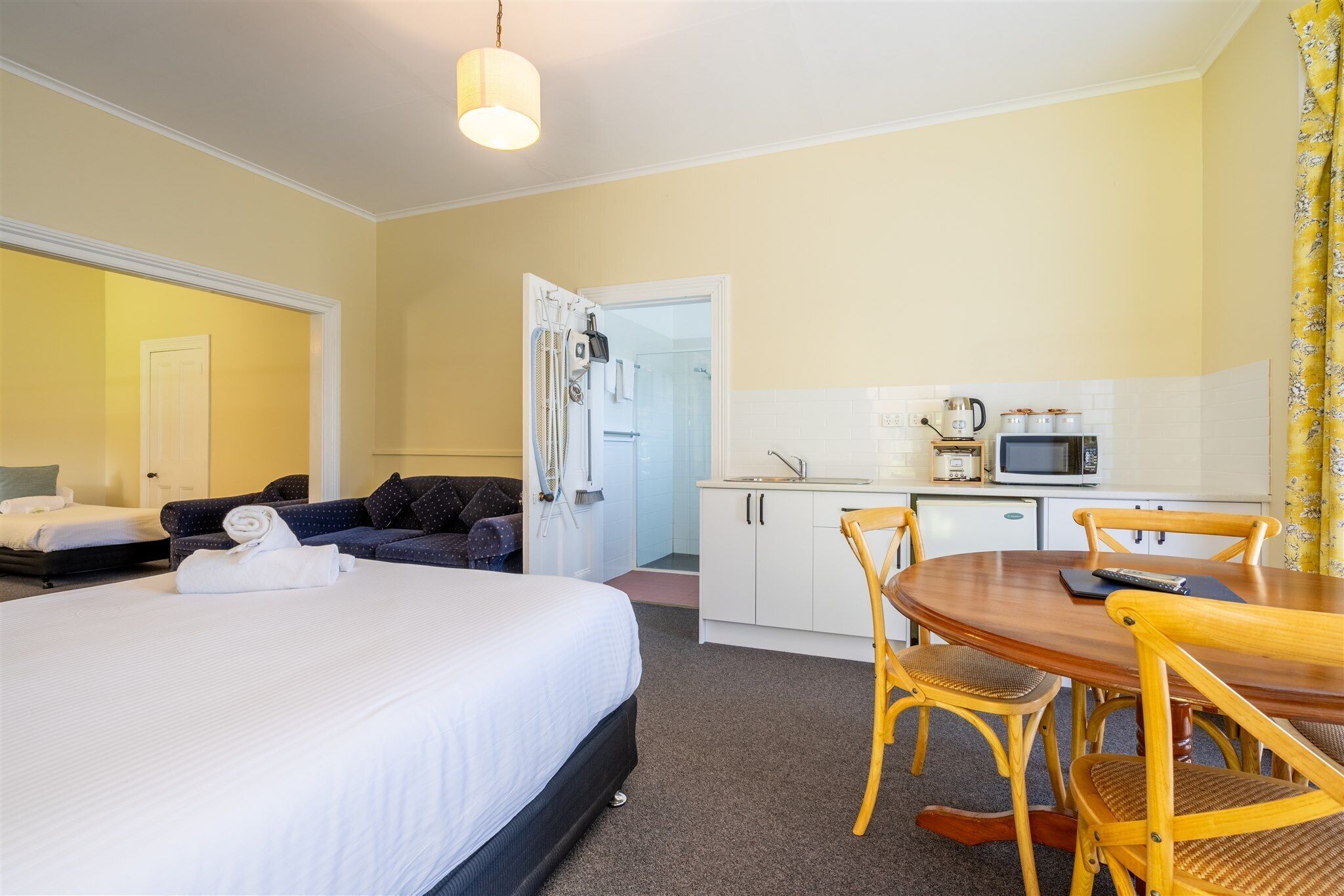 A hotel room with a bed, a sofa, a dining table with chairs, a kitchenette, and a shower area.
