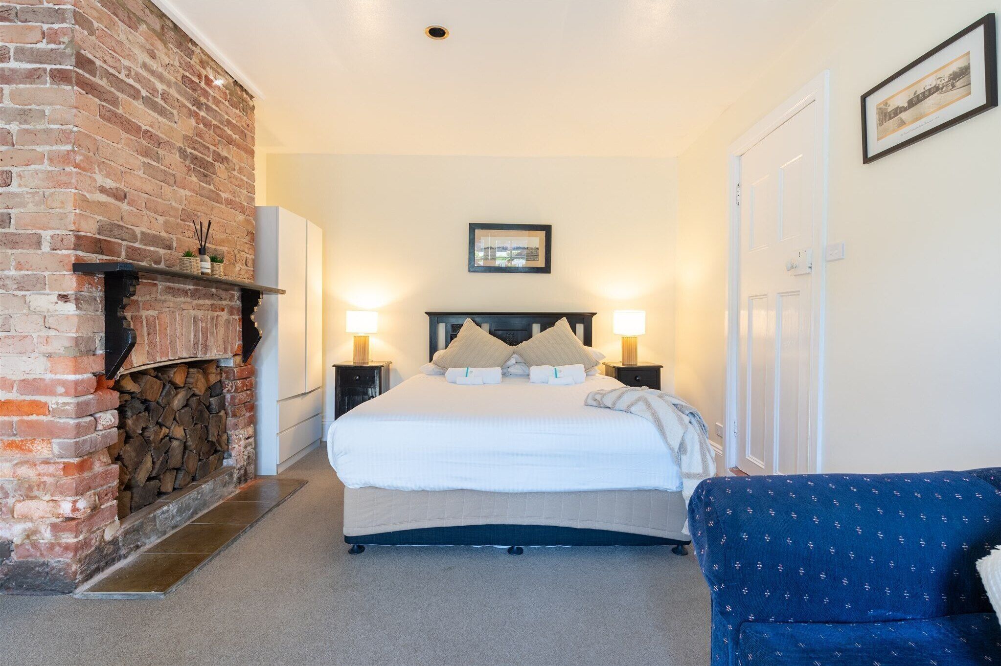 A bedroom with a brick fireplace, a bed with white bedding, two bedside lamps, and a framed picture on the wall.