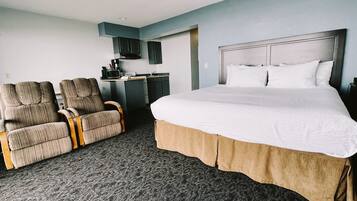 A hotel room with a large bed, two armchairs, a kitchenette, and a gray carpet.