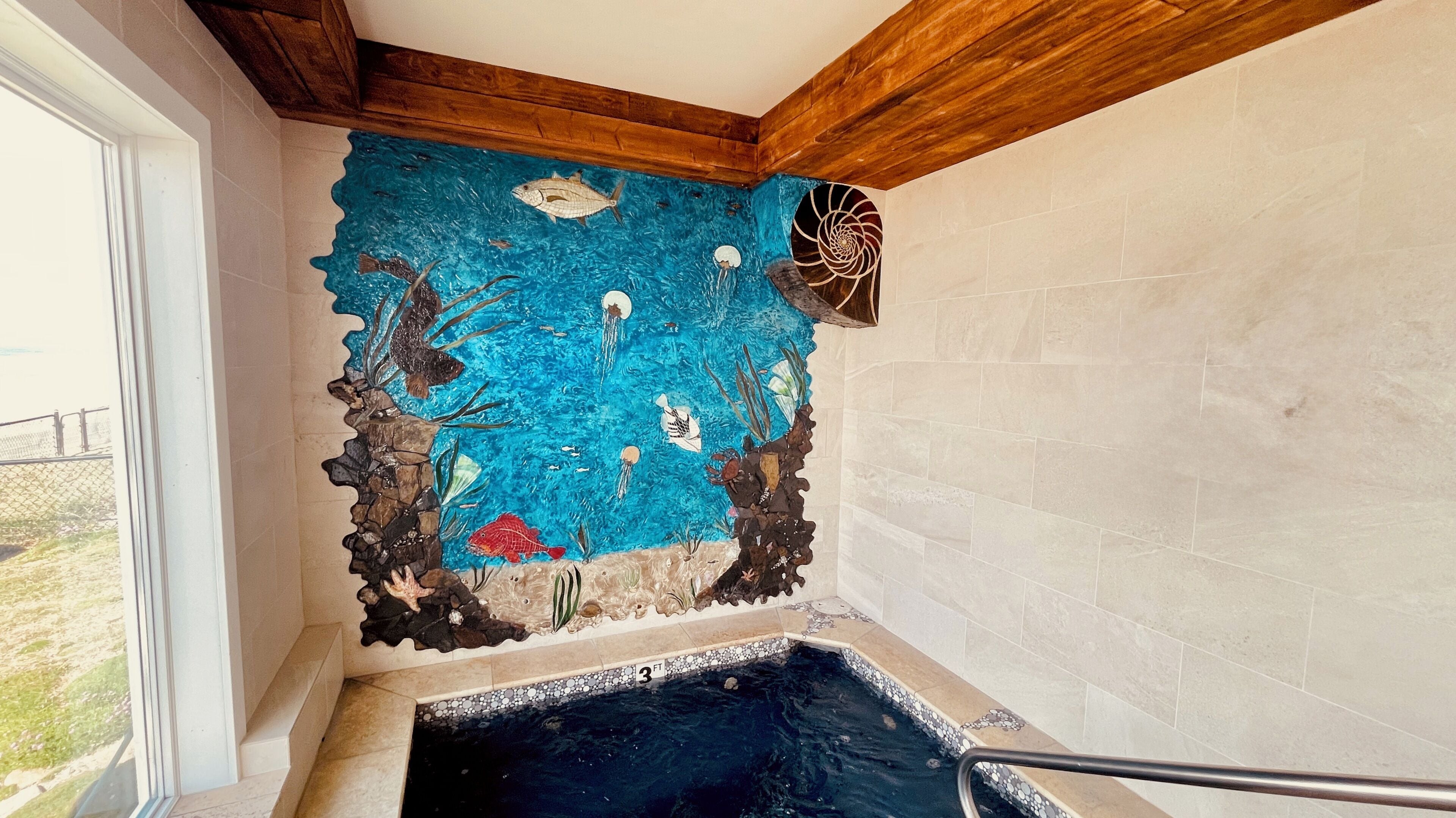 Esquina de una habitación con un mural azul que representa escenas submarinas, una pared de azulejos y una pequeña piscina.