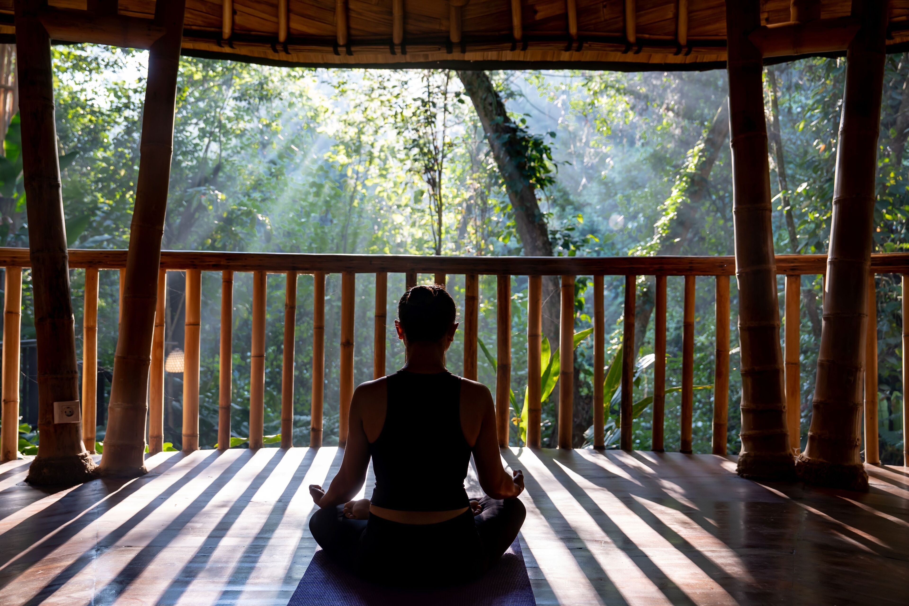 A person meditating on a wooden deck with bamboo pillars, overlooking a forested area.