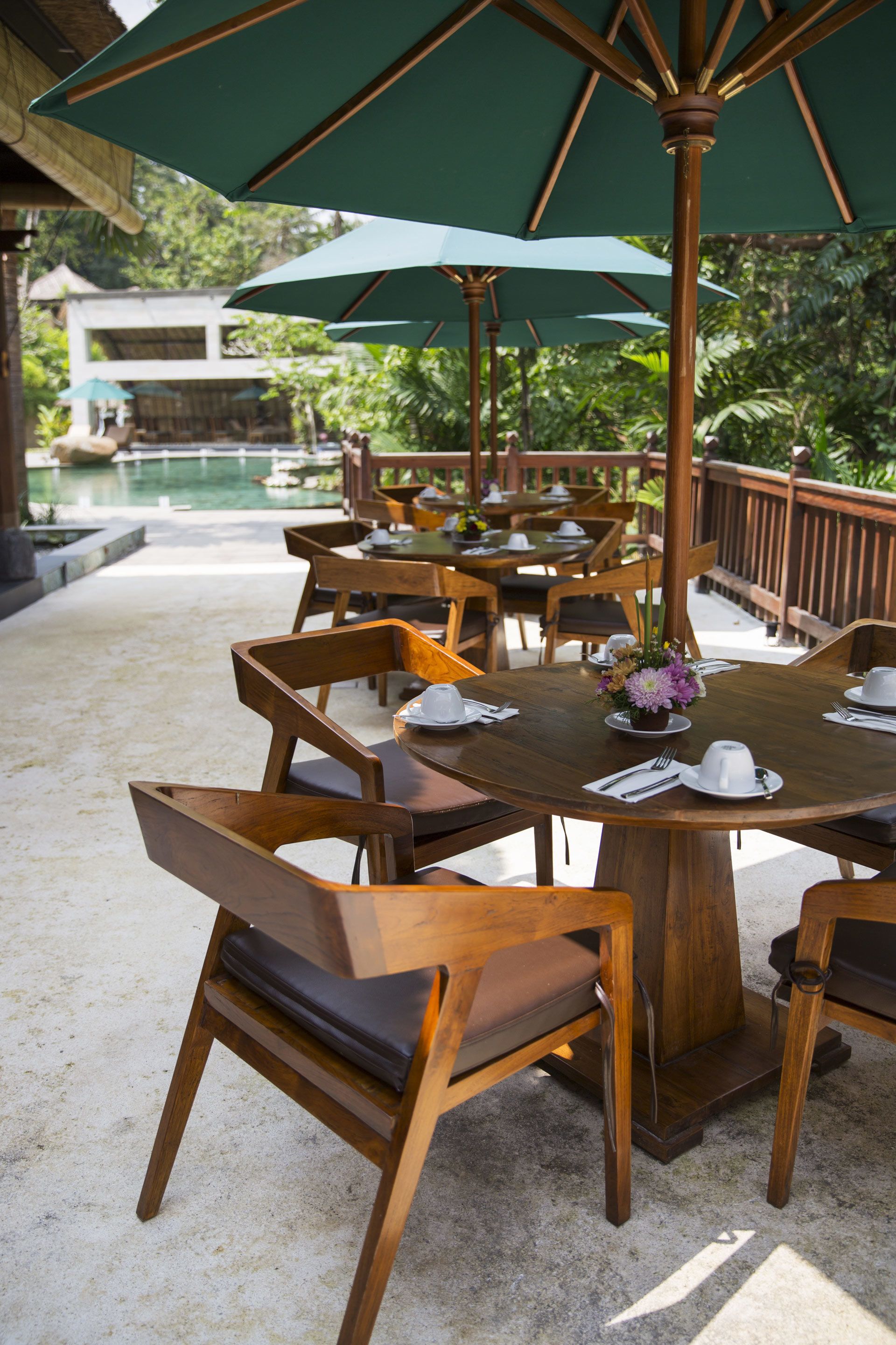 A wooden outdoor dining area with tables and chairs, surrounded by umbrellas and a pool in the background.