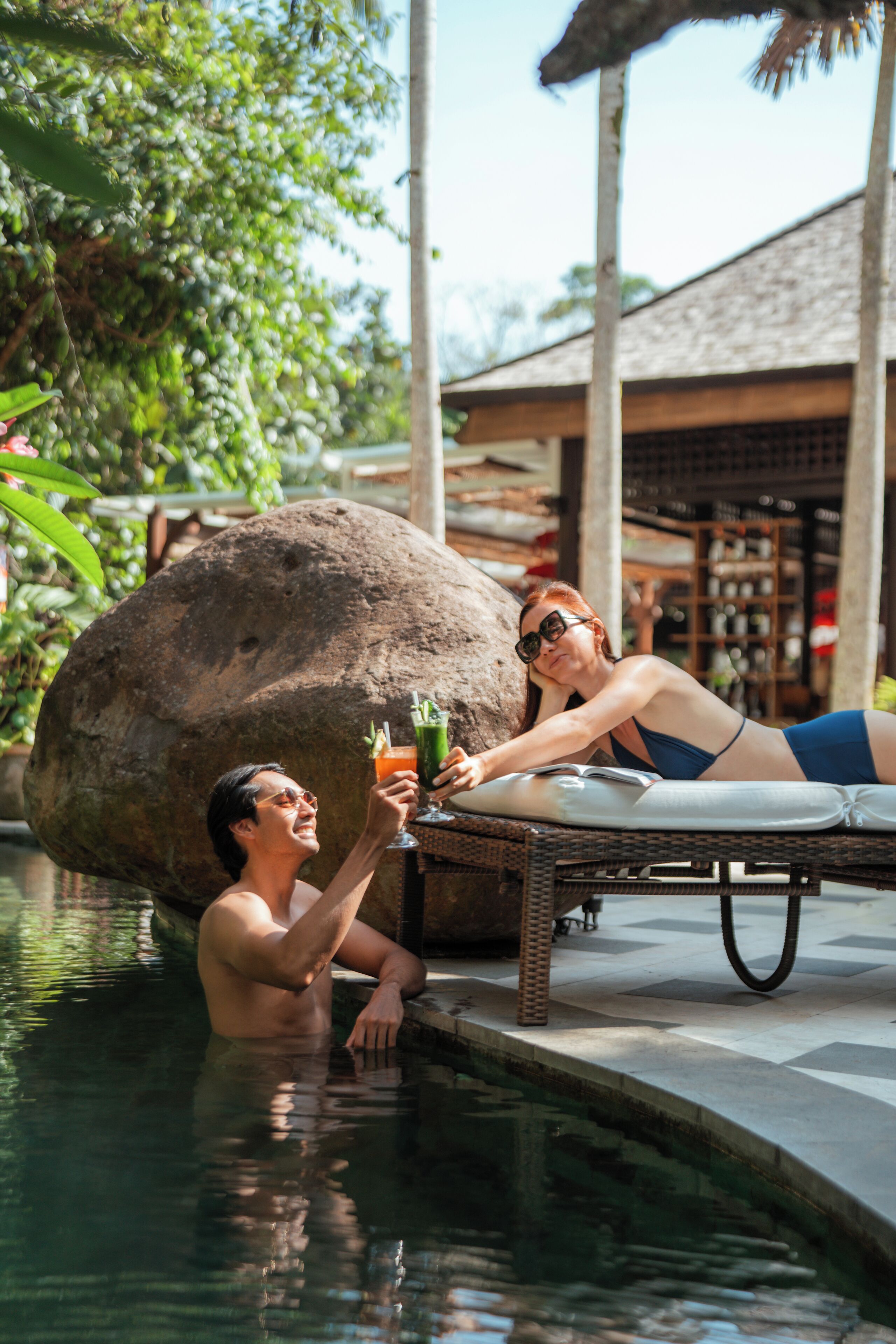 A couple relaxing by a poolside lounge chair, with a large rock and a bar area in the background.