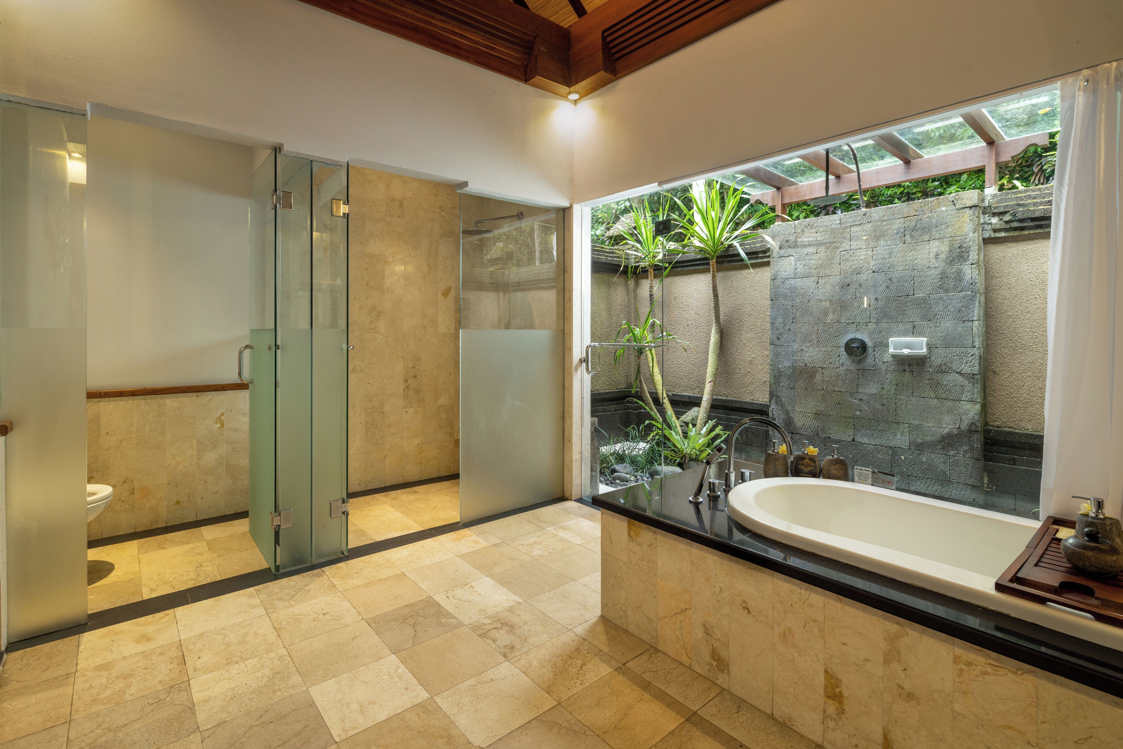 A modern bathroom with a glass shower enclosure, a freestanding bathtub, and a view of a garden through a large window.
