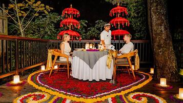 Un área de comedor al aire libre con una mesa lista para dos personas, rodeada de un colorido tapiz floral y sombrillas rojas.
